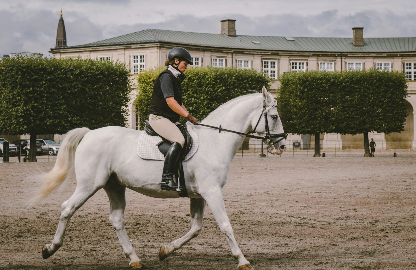 Jinete con casco hace trotar a un caballo blanco en una arena junto a un edificio histórico.